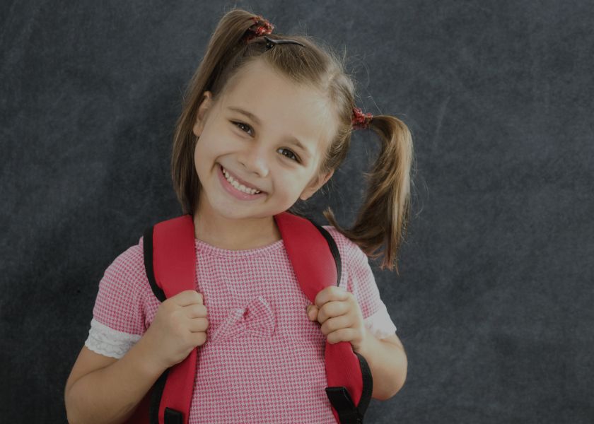 An elementary student smiling, with pig-tails, a red gingham shirt and a red backpack over her shoulders