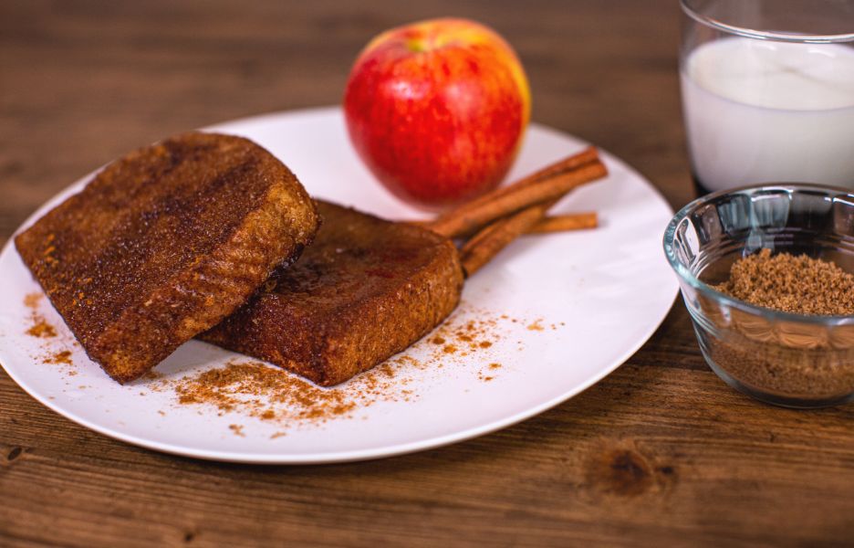 Cinnamon French Toast plated with dusted cinnamon and an apple, accompanied by a glass of milk and a serving dish of extra cinnamon