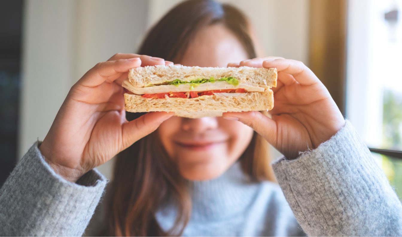 A young girl in a sweater, smiling, while holding up a turkey sandwich with lettuce and tomato in front of her eyes